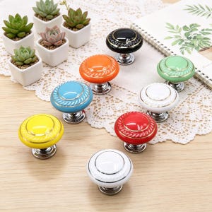 Könnte beinhalten: Eine Sammlung farbenfroher Keramik-Schrankknöpfe mit silberfarbenen Sockeln. Die Knöpfe sind in verschiedenen Farben erhältlich, darunter Gelb, Rot, Blau, Orange, Grün, Schwarz und Weiß, mit goldenen oder silbernen Details. Die Knöpfe sind auf einer Holzoberfläche angeordnet.