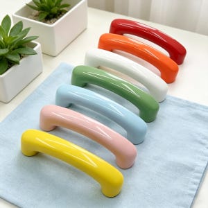 Könnte beinhalten: Sieben bunte Schrankgriffe in Regenbogenfarben: Rot, Orange, Weiß, Grün, Hellblau, Rosa und Gelb. Die Griffe sind auf einem hellblauen Tuch angeordnet. Ergonomisches Design für einfache Handhabung.