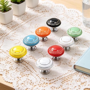 May include: A collection of eight colorful ceramic cabinet knobs. The knobs are in various colors, including yellow, blue, orange, red, white, black, and green. Each knob has a decorative design and a silver-colored base.