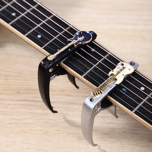 Op de afbeelding: Twee gitaarcapo's op een gitaarhals. Eén capo is zwart met een blauw gitaarontwerp, de andere zilver met een wit gitaarontwerp. De capo's zijn van metaal en hebben een veermechanisme.