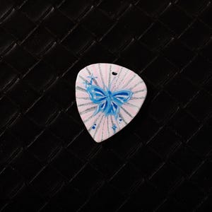 Può includere: Un plettro bianco con un disegno dipinto raffigurante un fiocco blu e linee radiali. Il plettro ha un foro nella parte superiore ed è posto su uno sfondo nero strutturato.