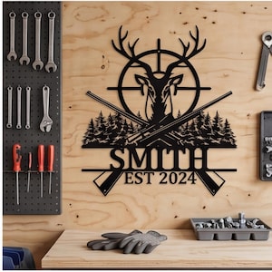 May include: A black metal wall sign featuring a deer head silhouette inside a circular crosshair target, crossed rifles, and a forest landscape. The sign includes the text SMITH and EST 2024 in a bold, capitalized font centered below the graphic.