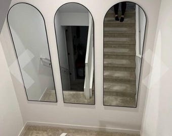 May include: Three arched mirrors with black frames are mounted on a white wall. The mirrors reflect a staircase and hallway. The mirrors are different sizes and are arranged in a row.