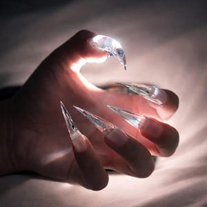 May include: A hand with long, clear acrylic nails, each shaped into sharp points. The nails are translucent, catching the light and creating a dramatic effect. The background is softly lit, emphasizing the nails' unique design.