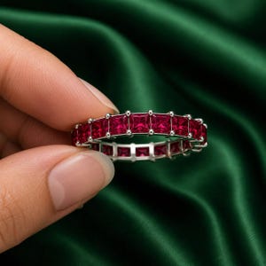 Könnte beinhalten: Ein silberner Eternity-Ring mit einer durchgehenden Reihe quadratisch geschliffener, tiefroter Rubine in einer Krappenfassung. Der Ring besteht aus einem Metallrahmen, der jeden Edelstein sicher hält und ein gleichmäßiges, rundes Design bildet.