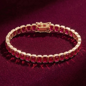 Könnte beinhalten: Ein goldfarbenes Armband mit rechteckigen, roten Edelsteinen. Das Armband liegt auf einer dunkelroten Samtoberfläche. Der Verschluss ist sichtbar, die Edelsteine sind gleichmäßig angeordnet.