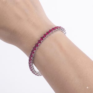 Könnte beinhalten: Ein Tennisarmband mit einer durchgehenden Reihe runder, leuchtend roter Edelsteine, die in ein poliertes, silberfarbenes Metallband gefasst sind. Das Armband umschließt das Handgelenk und zeigt ein einheitliches Design, bei dem jeder Stein in einer Krappenfassung sitzt.