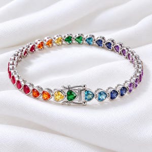 May include: A silver-toned tennis bracelet featuring a continuous row of heart-shaped gemstones in a vibrant rainbow spectrum, including red, orange, yellow, green, blue, and purple stones, secured with a sturdy metal box clasp closure.