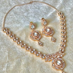 K&ouml;nnte beinhalten: Ein ros&eacute;goldenes Halsketten- und Ohrring-Set. Die Halskette und die Ohrringe sind mit klaren, runden und baguettef&ouml;rmigen Steinen besetzt. Die Ohrringe haben einen gro&szlig;en, klaren Stein in der Mitte und einen tropfenf&ouml;rmigen Anh&auml;nger.