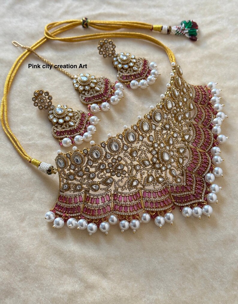 May include: A gold-toned necklace and earring set with a matching headpiece. The jewelry features clear and pink gemstones, and white pearls. The necklace has a wide, ornate design with a pink border. The text "Pink city creation Art" is visible.