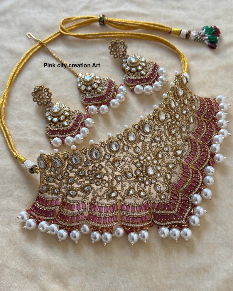 May include: A gold-toned necklace and earring set with a matching headpiece. The jewelry features clear and pink gemstones, and white pearls. The text "Pink city creation Art" is visible on the image.