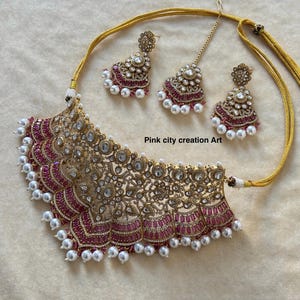 May include: A gold-toned necklace and earring set with a matching headpiece. The necklace features a detailed, ornate design with red and clear stones, and white pearls. The earrings and headpiece mirror the necklace's design. The text "Pink city creation Art" is visible.