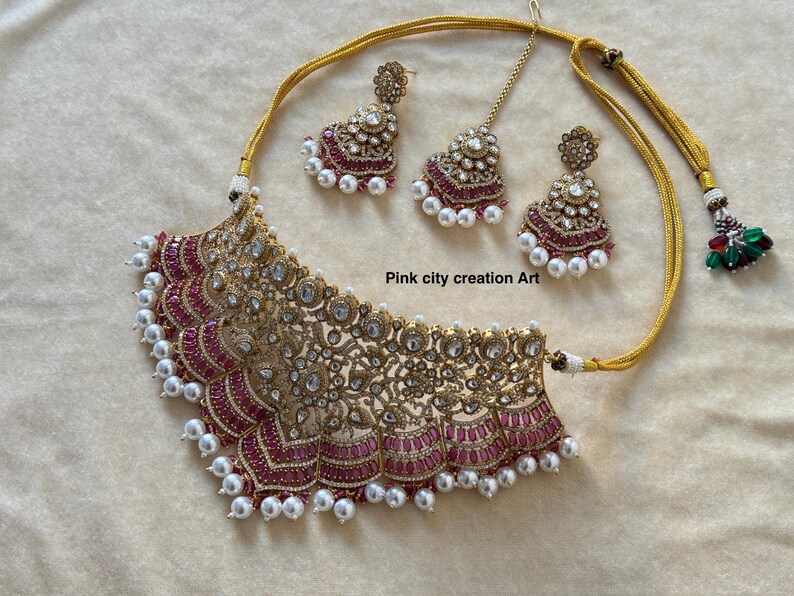 May include: A gold-toned necklace and earring set with a matching headpiece. The necklace features a detailed, ornate design with red and clear stones, and white pearls. The earrings and headpiece mirror the necklace's design. The text "Pink city creation Art" is visible.