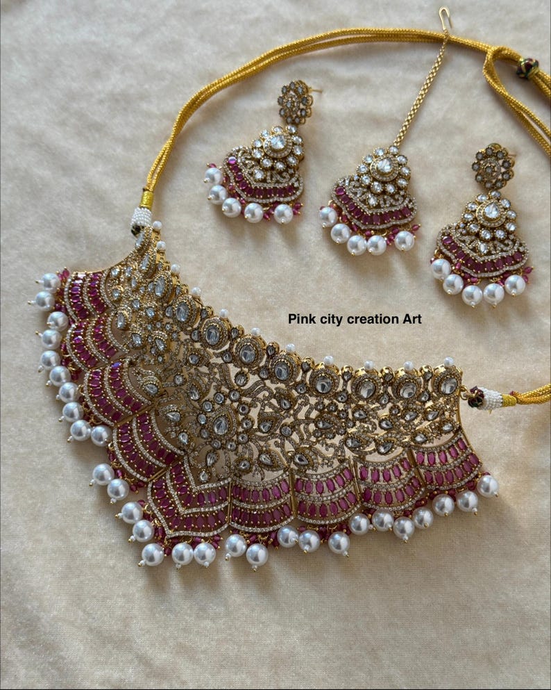 May include: A gold-toned necklace and earring set with a matching headpiece. The necklace and earrings feature intricate detailing with clear and pink gemstones, and white pearls. The text "Pink city creation Art" is visible.