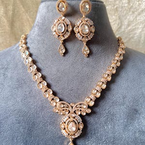 K&ouml;nnte beinhalten: Ein goldfarbenes Schmuckset mit Halskette und passenden Ohrringen. Die Halskette hat einen zentralen Anh&auml;nger mit einem gro&szlig;en, klaren Stein und einem tropfenf&ouml;rmigen Anh&auml;nger. Die Ohrringe spiegeln das Anh&auml;ngerdesign wider und schaffen einen stimmigen Look.