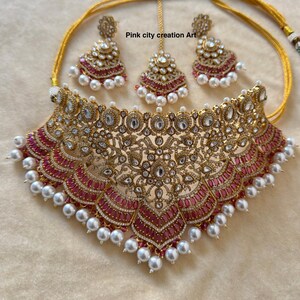 May include: A gold-toned jewelry set featuring a necklace, earrings, and a headpiece. The necklace and earrings have a floral design with red and clear stones, and white pearls. The headpiece is a smaller version of the earrings.