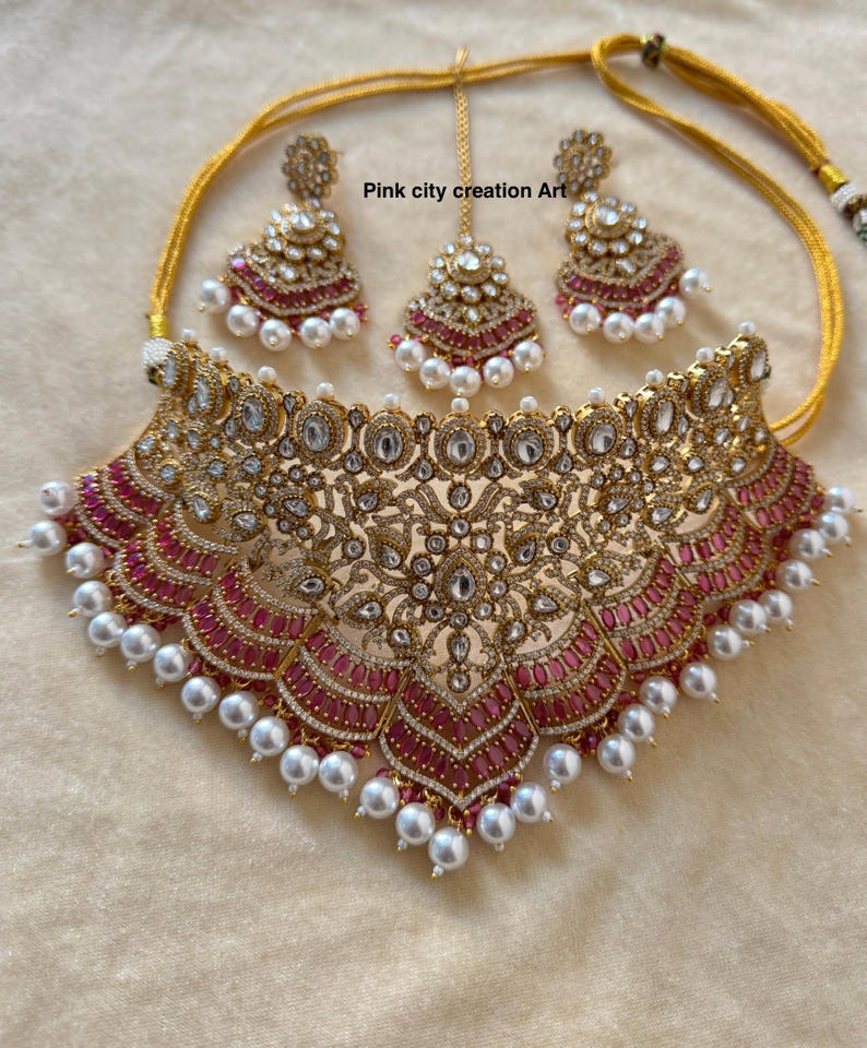 May include: A gold-toned jewelry set featuring a necklace, earrings, and a headpiece. The necklace and earrings have a floral design with red and clear stones, and white pearls. The headpiece is a smaller version of the earrings.