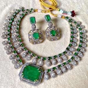 Könnte beinhalten: Ein zweiteiliges Schmuckset mit Halskette und Ohrringen. Die Halskette und Ohrringe sind mit großen, smaragdgrünen Edelsteinen besetzt, die von kleineren, klaren Steinen umgeben sind. Die Halskette hat einen großen, smaragdgeschliffenen Mittelstein.
