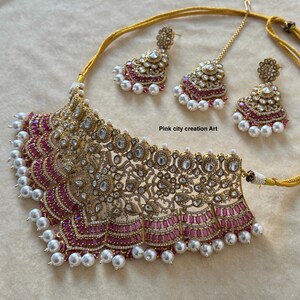 May include: A gold-toned necklace, earrings, and headpiece set. The necklace features a wide, ornate design with pink and clear stones, and white pearls. The earrings and headpiece mirror the necklace's design. The text "Pink city creation Art" is visible.