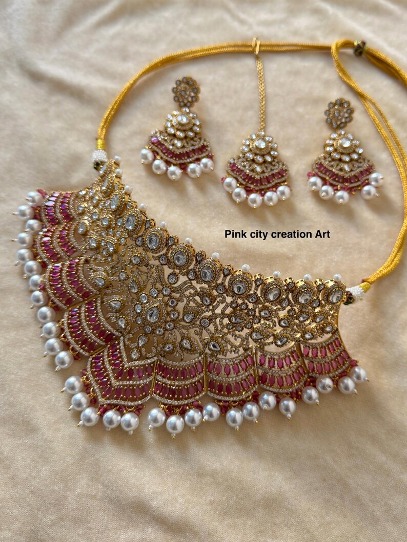 May include: Gold-toned necklace and earring set with ruby-colored accents and white pearls. The necklace features intricate filigree and sparkling clear stones. The set includes matching earrings and a headpiece. Text on the image reads "Pink city creation Art."