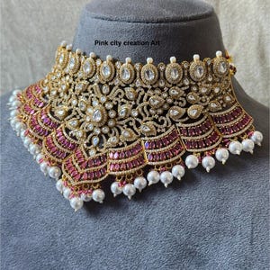 May include: A gold-toned statement necklace with intricate detailing, featuring clear and ruby-colored stones, and dangling white pearls. The necklace is displayed on a gray mannequin head. Text on the image reads "Pink city creation Art".