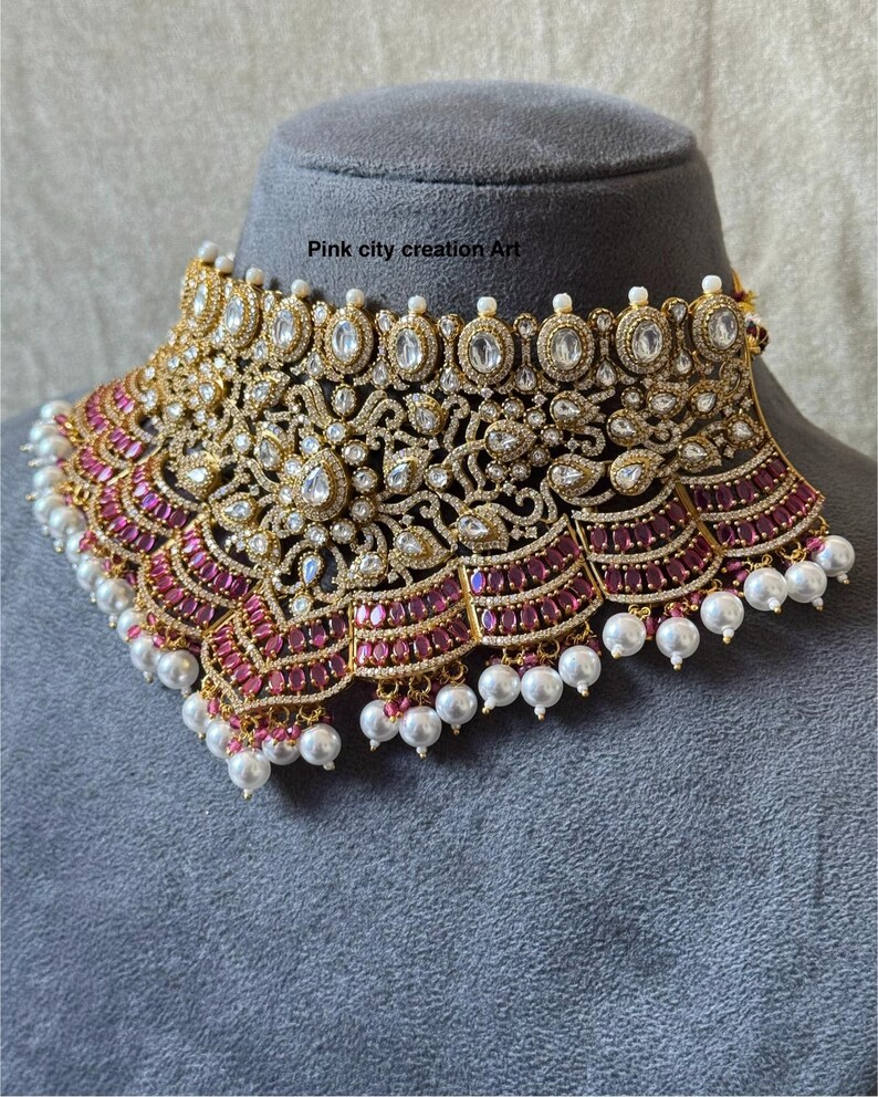 May include: A gold-toned statement necklace with intricate detailing, featuring clear and ruby-colored stones, and dangling white pearls. The necklace is displayed on a gray mannequin head. Text on the image reads "Pink city creation Art".