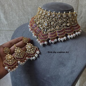 May include: A gold-toned necklace and matching earrings set. The necklace is ornate with clear and ruby-colored stones, and white pearls. The earrings have a similar design. The text "Pink city creation Art" is visible.