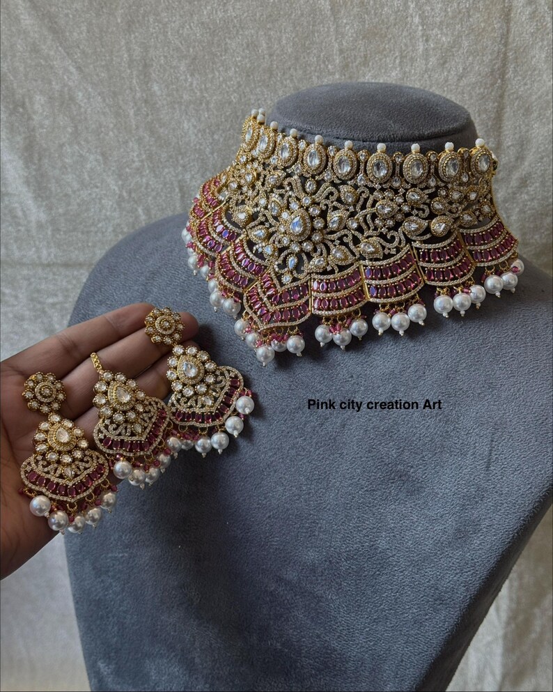 May include: A gold-toned necklace and matching earrings set. The necklace is ornate with clear and ruby-colored stones, and white pearls. The earrings have a similar design. The text "Pink city creation Art" is visible.