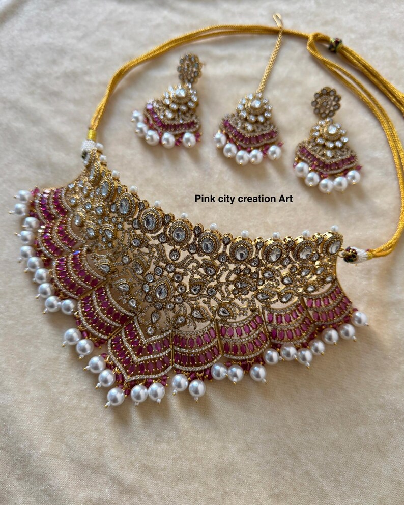 May include: A gold-toned necklace and earring set with a matching headpiece. The jewelry features intricate filigree designs, clear gemstones, and pink accents. The necklace and earrings are adorned with white pearls. The text "Pink city creation Art" is visible.