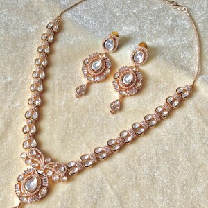 K&ouml;nnte beinhalten: Ein ros&eacute;goldenes Halsketten- und Ohrring-Set. Die Halskette hat einen zentralen Anh&auml;nger mit einem gro&szlig;en, klaren Stein und kleinere Steine. Die Ohrringe haben ein &auml;hnliches Design. Das Schmuckst&uuml;ck liegt auf einer cremefarbenen Oberfl&auml;che.