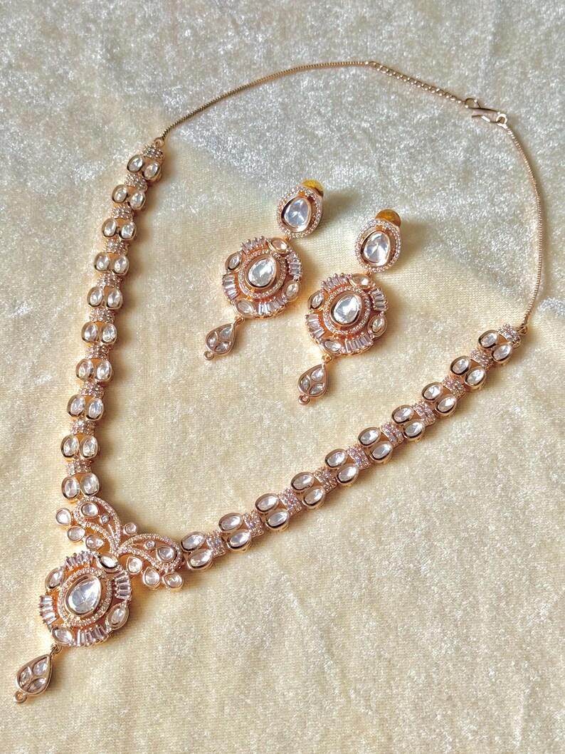 K&ouml;nnte beinhalten: Ein ros&eacute;goldenes Halsketten- und Ohrring-Set. Die Halskette hat einen zentralen Anh&auml;nger mit einem gro&szlig;en, klaren Stein und kleinere Steine. Die Ohrringe haben ein &auml;hnliches Design. Das Schmuckst&uuml;ck liegt auf einer cremefarbenen Oberfl&auml;che.