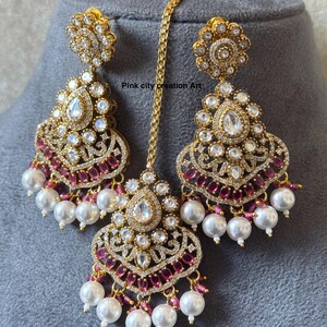 May include: Gold-toned, ornate jewelry set featuring a pair of drop earrings and a headpiece. The earrings and headpiece are adorned with clear and ruby-colored stones, and white pearl drops. The text "Pink city creation Art" is visible.
