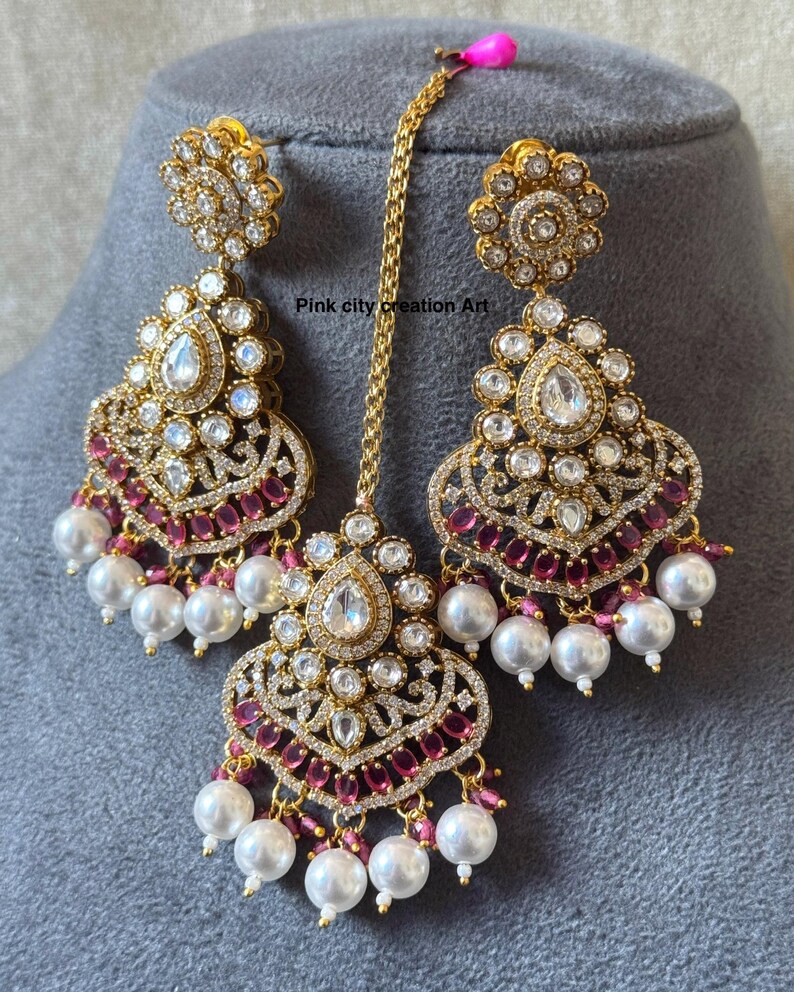 May include: Gold-toned, ornate jewelry set featuring a pair of drop earrings and a headpiece. The earrings and headpiece are adorned with clear and ruby-colored stones, and white pearl drops. The text "Pink city creation Art" is visible.