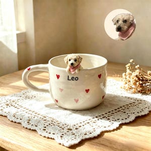 May include: A white ceramic mug featuring a small, sculpted dog figurine with a pink bandana peering over the rim. The mug is decorated with small red hearts and the name Leo printed in black text. A circular inset shows the dog wearing a bandana.