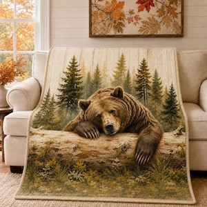 May include: A rectangular throw blanket featuring a printed image of a brown bear resting its head and paws on a fallen log. The background displays a dense forest of evergreen trees with a light wood-grain texture overlaying the entire design.