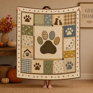 Könnte beinhalten: Eine gesteppte Wohndecke mit einem großen Pfotenabdruck in der Mitte, umgeben von Patchwork-Quadraten in Blau-, Grün- und Beigetönen. Das Design umfasst Karomuster, Vichy-Karo, Blumenmotive, Hunde- und Katzensilhouetten sowie kleine Pfoten.