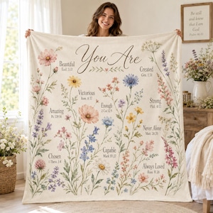Peut inclure: Une couverture en polaire crème ornée d'un motif floral avec diverses fleurs sauvages et le texte "You Are" suivi de mots inspirants et de versets bibliques, notamment Beautiful, Created, Victorious, Enough, Strong, Amazing, Never Alone, Chosen, Capable et Always Loved.
