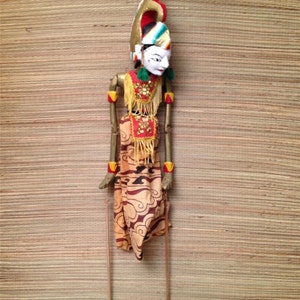 May include: A traditional Indonesian puppet with a colorful headdress and a painted face. The puppet is wearing a patterned orange and brown robe and is attached to two wooden sticks.