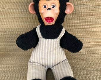 May include: A black plush monkey wearing a striped overall and a red and white striped hat. The monkey has a wide smile and large, round eyes.