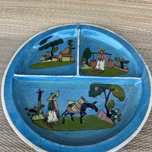 May include: A divided ceramic plate with a vibrant blue glaze. The plate features three sections, each with a hand-painted scene of figures, trees, and buildings in a folk art style. The rim is a lighter shade of blue.