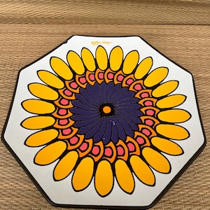 May include: A white octagonal plate with a colorful sunflower design. The center of the sunflower is a blue and pink spiral with a yellow center. The sunflower petals are yellow with orange outlines. The plate has the text "Peter Max" in black lettering.