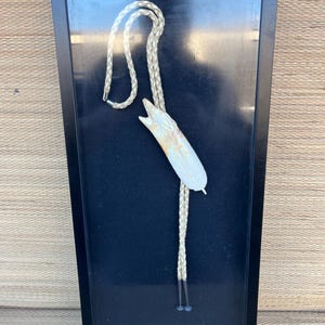 May include: A framed bolo tie with a white feather and a braided leather cord. The bolo tie is displayed against a black background.