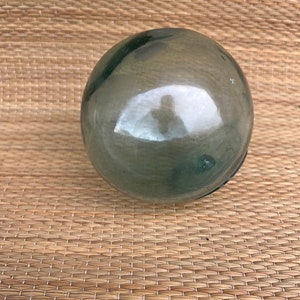 May include: A large, round, clear glass buoy with a greenish tint. The buoy is sitting on a woven mat.