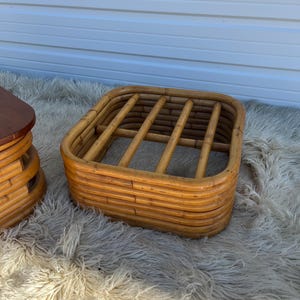 May include: A woven rattan ottoman with a square top and a rounded frame. The ottoman is made of natural brown rattan and has a woven pattern.