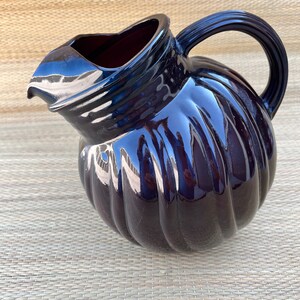 May include: A dark blue ceramic pitcher with a ribbed pumpkin-like design. The pitcher has a curved handle and a spout for pouring. The glossy finish reflects light, highlighting the curves and details of the pitcher. A decorative kitchenware item.