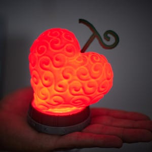 Peut inclure: Lampe rouge en forme de cœur avec un motif tourbillonnant, ressemblant à un fruit stylisé. La lampe a une tige verte et repose sur une base grise et rouge. La lampe est tenue dans la main d'une personne.
