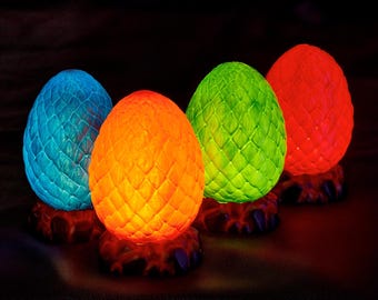 May include: Four glowing, egg-shaped decorative lights in blue, orange, green, and red. Each egg has a textured, scale-like pattern and sits on a dark, rocky base. The lights emit a warm glow.