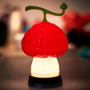 May include: A decorative mushroom-shaped lamp with a bright red cap and a white, swirling pattern base. The lamp has a green stem and a small green top. The base is black. The lamp is lit, emitting a soft glow.