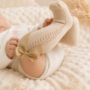 Könnte beinhalten: Ein Baby trägt beige kniehohe Socken mit einer dekorativen Schleife. Die Socken haben ein zartes Lochmuster. Das Baby liegt auf einer weichen, strukturierten Decke. Die Farbpalette ist neutral, mit Beige- und Cremetönen.