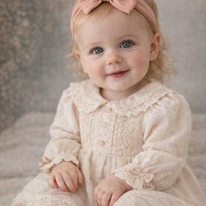 Könnte beinhalten: Ein lächelndes Baby mit blauen Augen trägt ein hellbeige Kleid mit Spitzenbesatz und ein passendes rosa Stirnband mit Schleife. Das Kleid hat lange Ärmel und eine Knopfleiste. Das Baby trägt auch rosa Socken mit Schleifen.